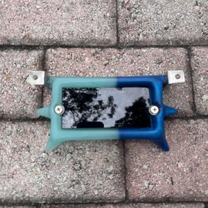 May include: A small rectangular device with a screen, framed by a two-tone blue and teal plastic casing. The device has metal brackets on each end and is secured with screws. The screen reflects the surrounding environment.