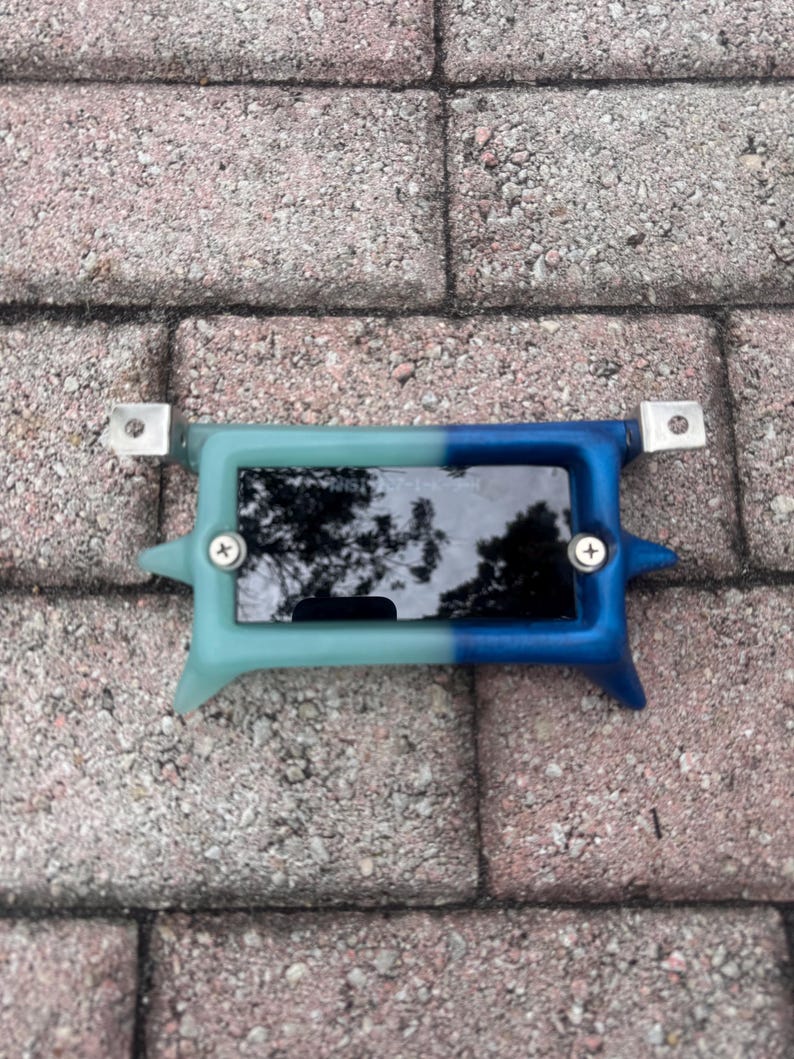 May include: A small rectangular device with a screen, framed by a two-tone blue and teal plastic casing. The device has metal brackets on each end and is secured with screws. The screen reflects the surrounding environment.