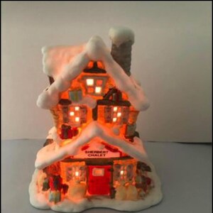 Op de afbeelding: Een wit, met sneeuw bedekt, miniatuur peperkoekhuis met een rode deur en de tekst "Sherbert Chalet" op de voorkant. Het huis is verlicht met warme, gele lampjes.