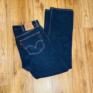 May include: Dark blue denim jeans with a classic five-pocket design. The jeans feature a red and white label on the back pocket and a small red tag. The jeans are folded on a wooden surface.
