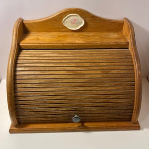 Può includere: Un portapane in legno con anta a serrandina e un piccolo pomello argentato. L'unità presenta un ripiano aperto nella parte superiore, decorato con una piccola placca ovale che mostra un motivo floreale con fiori rosa e blu.