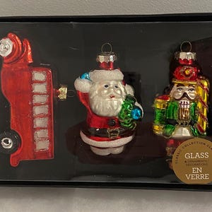 May include: A set of four glass Christmas ornaments in a black box. The ornaments include a red bus, Santa Claus, and a nutcracker. The ornaments have glitter accents and are in a decorative box. The box has a gold label that says "Glass" and "En Verre".