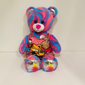 Può includere: Un orsacchiotto di peluche con un motivo a spirale rosa, blu e viola. L'orso indossa un grembiule rosa con un design a cartoni animati e ha accenti argentati sulle zampe e sulle orecchie. Ha un naso rosa ed è seduto in posizione eretta. Giocattolo per bambini.