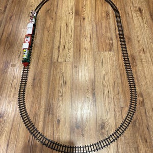 May include: A toy train set consisting of a green, red, and white locomotive followed by three train carriages, positioned on an oval-shaped black plastic track. The train features a festive colour scheme with small figures visible on the rear carriage.