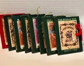 May include: A collection of Christmas ornaments shaped like miniature books. The ornaments have a dark green cover with gold lettering and illustrations. Each ornament has a red ribbon for hanging. The text "Old World Toy Collection" is visible.