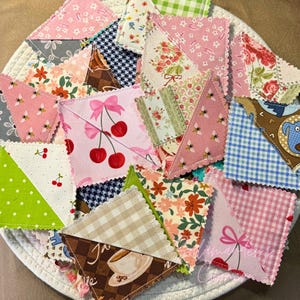 May include: A collection of handmade fabric bookmarks in various patterns and colors. Designs include cherries, bees, floral prints, polka dots, and gingham. Each bookmark is a unique patchwork of different fabrics, with scalloped edges.