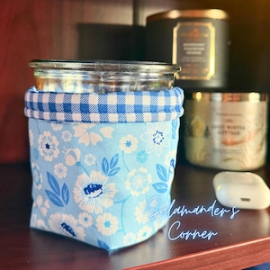 May include: A glass jar with a blue floral fabric sleeve. The sleeve features white flowers and blue leaves on a light blue background. The top of the sleeve has a blue and white gingham pattern. The text "Salamanders Corner" is visible.