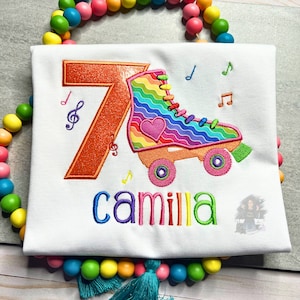 May include: A white t-shirt with a rainbow roller skate and the number 7 embroidered on it. The name "camilla" is embroidered below the roller skate.