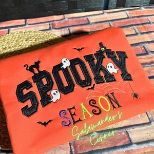 May include: An orange sweatshirt featuring the black glittery text SPOOKY SEASON. The design includes embroidered white ghosts, a black cat, bats, a spider, a spiderweb, and a witch hat. The text Salamanders Corner is embroidered in light green.