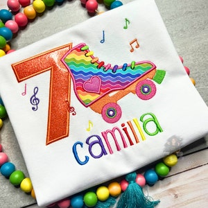 May include: A white t-shirt with an embroidered design of a rainbow roller skate with a heart and the number 7. The name "camilla" is embroidered below the roller skate.