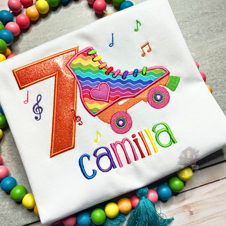 May include: A white t-shirt with an embroidered design of a rainbow roller skate with a heart and the number 7. The name "camilla" is embroidered below the roller skate.