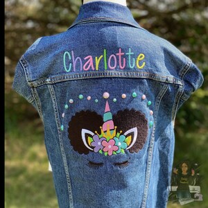 May include: A sleeveless denim jacket with the name "Charlotte" embroidered in colorful thread across the upper back. Below the name is a unicorn design with a brown afro, a pink horn, and floral accents. The jacket is decorated with iridescent beads.