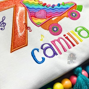 May include: A white shirt with a rainbow roller skate design and the number 7 embroidered on it. The name "camilla" is also embroidered on the shirt in rainbow colors.