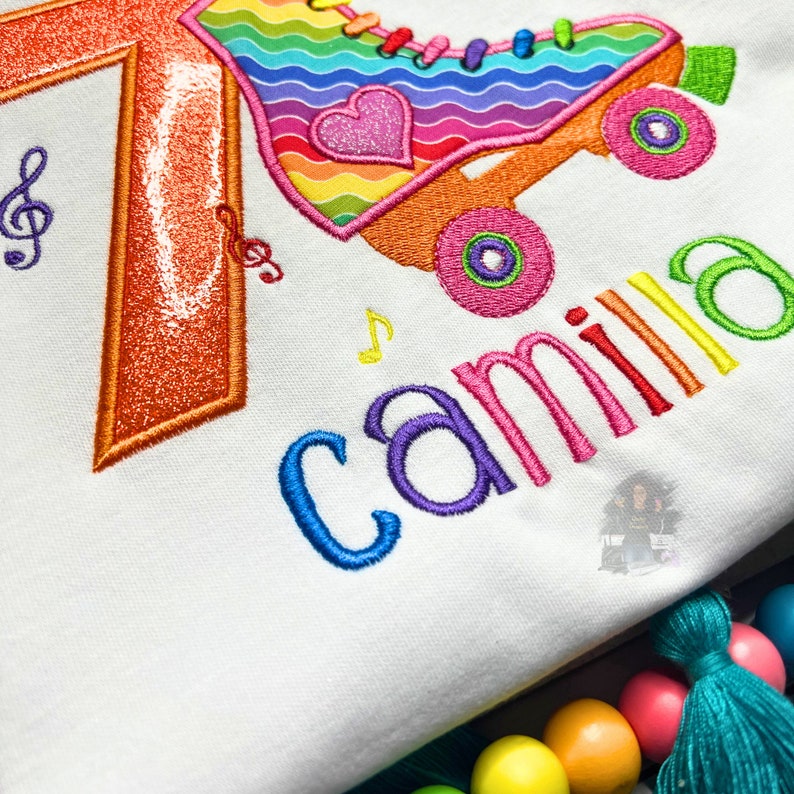 May include: A white shirt with a rainbow roller skate design and the number 7 embroidered on it. The name "camilla" is also embroidered on the shirt in rainbow colors.