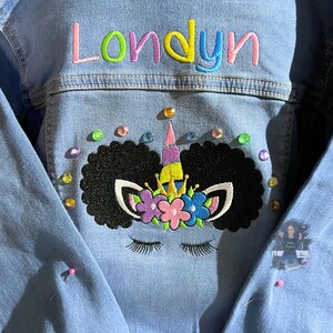 May include: A blue denim jacket with the name "Londyn" embroidered on the back. The jacket features a black and white unicorn with a rainbow horn and a crown of flowers. The unicorn is surrounded by colorful rhinestones.