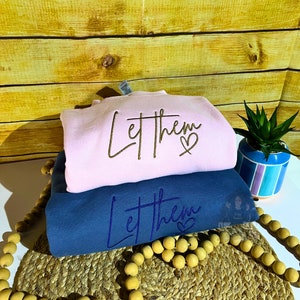 May include: Two sweatshirts stacked on top of each other. The top sweatshirt is pink and says "Let them" with a heart. The bottom sweatshirt is blue and says "Let them" with a heart.