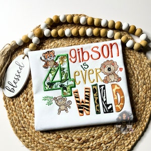 May include: White onesie with a green number four, surrounded by jungle animals and foliage. The text "Gibson is ever wild" is embroidered in orange and brown.