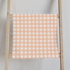 Op de afbeelding: Een perzikkleurige gingham stof met een wit geruit patroon. De stof is over een houten ladder gelegd.