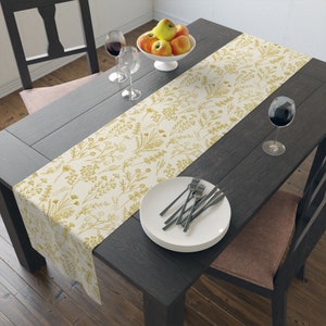 May include: A white table runner with a gold floral pattern. The runner is laid on a dark wood table with a bowl of apples, two wine glasses, and a set of white plates and silverware.