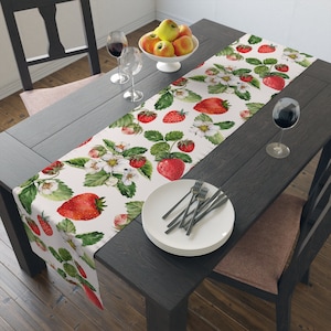 Peut inclure: Un chemin de table blanc avec un motif répété de fraises rouges et de fleurs blanches. Le chemin de table est posé sur une table en bois foncé avec un bol de pommes, deux verres à vin et un couvert.