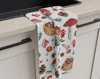 Op de afbeelding: Een witte keukendoek met een herhalend patroon van rode en bruine paddenstoelen, dennenappels en groene bladeren. De handdoek hangt aan een witte keukenkastdeur.