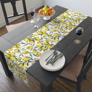 May include: A white table runner with a vibrant lemon and leaf pattern. The runner is placed on a dark wooden table, complemented by a white bowl of apples, wine glasses, and a plate with forks. The design features yellow lemons and green leaves.