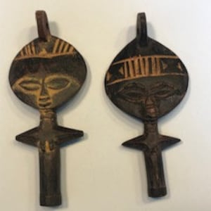 May include: Two wooden pendants shaped like a stylised human figure. The pendants are painted in shades of brown, green and gold. The figures have a simple design with a round head, a small body and outstretched arms.