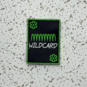 May include: A rectangular black patch with a green border. The patch features the word "WILDCARD" in white, with a green, wavy line above it. Green starburst designs are in the top left and bottom right corners.