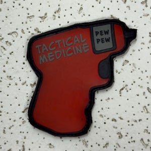May include: A red patch in the shape of a gun with the words "TACTICAL MEDICINE" and "PEW PEW" in grey lettering. The patch has a black border and is likely for a medical or tactical kit.