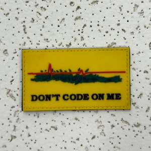 Könnte beinhalten: Ein rechteckiger gelber Aufnäher mit schwarzem Rand. Er zeigt eine rote Herzschlaglinie über einer grünen, stilisierten Baumlinie. Der Text "DON'T CODE ON ME" ist in schwarzen Großbuchstaben darunter gedruckt.