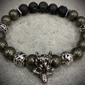 May include: A beaded bracelet with dark grey lava rock beads, metallic pyrite beads, and ornate silver-toned accent beads. It features a silver wolf head charm. The bracelet is strung on an elastic cord.