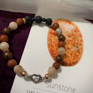 May include: A bracelet with a mix of brown, beige, and gold beads, including goldstone and lava rock. It has rose gold-coloured spacers and a heart-shaped clasp. The word "sunstone" is printed on a card, with the phrase "Take pleasure in the things you do".