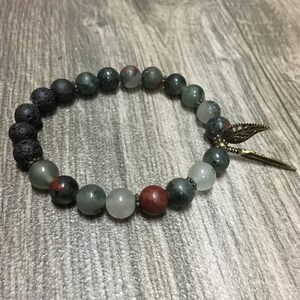 May include: A beaded bracelet featuring black lava rock, green and red jasper, and white moonstone beads. The bracelet has a gold-tone charm with a feather design.