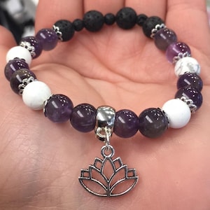 Op de afbeelding: Een armband met amethist, witte en zwarte lavabeads. Het heeft een zilveren lotusbloem bedel. De kralen worden gescheiden door zilverkleurige afstandhouders. De armband wordt op een hand getoond.
