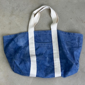 May include: A large blue tote bag with white canvas handles and accents. The bag has a rectangular shape with a pocket on the front. The bag is made of a durable material, perfect for carrying groceries or other items.