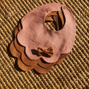 May include: A set of three layered bibs in pink and brown with a scalloped edge and a brown bow. The bibs are made of a soft, linen fabric.