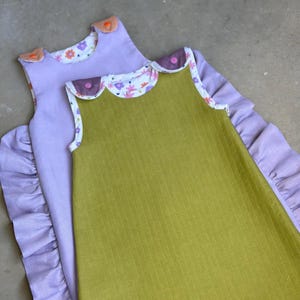 Könnte beinhalten: Zwei Babyschlafsäcke, einer ist lavendelfarben mit einem Blumendruck und der andere ist einfarbig olivgrün. Beide haben gekräuselte Ränder und sind aus weichem Stoff.