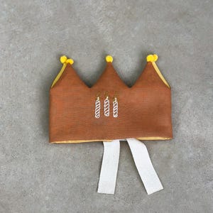 May include: A brown felt crown with yellow pom-poms and white ribbon ties. The crown has embroidered candles on it.