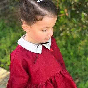 May include: A young child wearing a red dress with a white collar and a black bow. The child's hair is styled with a red patterned bow and a white barrette. The dress has a ruffled hem.