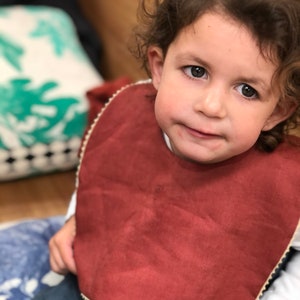 May include: A close-up of a young child wearing a rust-colored bib with a white trim. The bib is made of a soft, textured fabric. The child has brown hair and is looking directly at the camera.