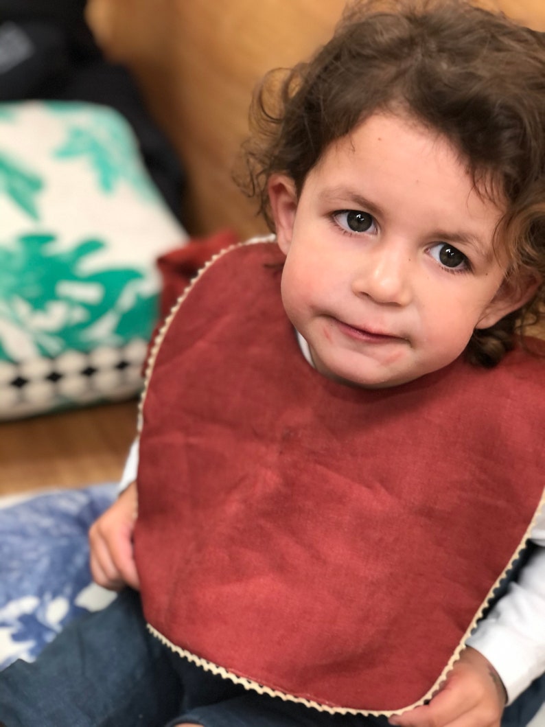 May include: A close-up of a young child wearing a rust-colored bib with a white trim. The bib is made of a soft, textured fabric. The child has brown hair and is looking directly at the camera.