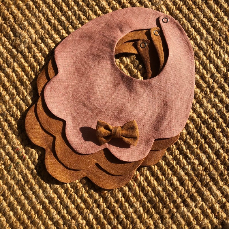 May include: A set of three layered bibs in pink and brown with a scalloped edge and a brown bow. The bibs are made of a soft, linen fabric.