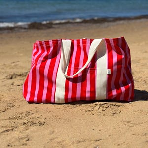 May include: A striped beach tote bag in red and pink, with white canvas handles and a pocket. The bag is on a sandy beach, with the sea visible in the background. The bag's dimensions are not visible.