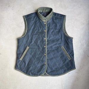 Può includere: Un gilet trapuntato blu scuro con finiture e colletto verde oliva. Il gilet presenta un motivo a rombi, una chiusura con bottoni e due tasche anteriori. Un capo di abbigliamento esterno classico.
