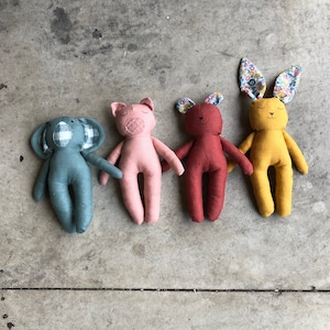 Puede incluir: Cuatro juguetes de peluche hechos a mano en diferentes colores y formas de animales: un elefante azul, un cerdo rosa, un oso rojo y un conejito amarillo. Los juguetes están sobre una superficie de hormigón gris.