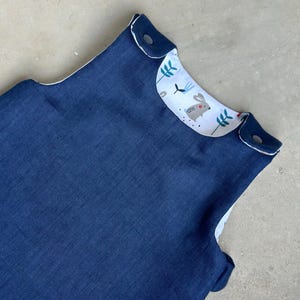 Puede incluir: Un saco de dormir azul marino para bebé con interior blanco. El cuello presenta una tela estampada con un conejito gris, flores azules y hojas verdes. El saco de dormir tiene cierres a presión.