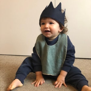 May include: A baby wearing a blue crown and a light blue bib sits on a brown carpet. The baby is wearing a dark blue sweatshirt and matching pants.