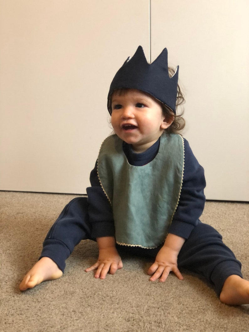 May include: A baby wearing a blue crown and a light blue bib sits on a brown carpet. The baby is wearing a dark blue sweatshirt and matching pants.