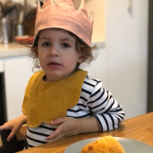 May include: A young child wearing a pink crown and a yellow bib sits at a table with a plate of food. The child is wearing a striped shirt and has curly brown hair.