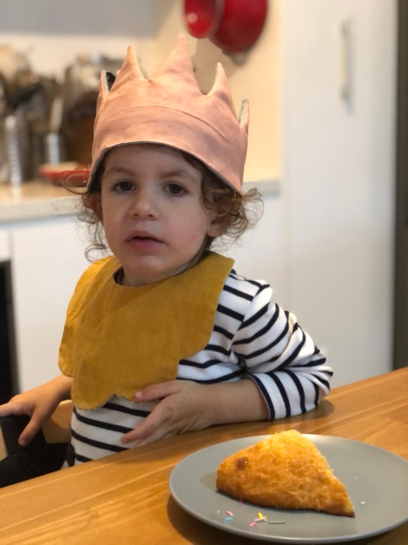May include: A young child wearing a pink crown and a yellow bib sits at a table with a plate of food. The child is wearing a striped shirt and has curly brown hair.